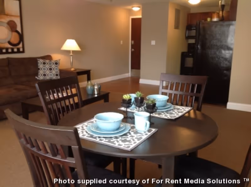 Open-plan living and dining area with a round dining table set with blue dishes, a sofa and lamp, and a kitchen with a black refrigerator in the background.