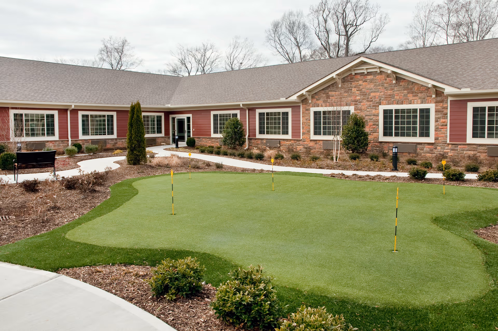 Outdoor courtyard area of a senior living facility with a putting green, small shrubs, a bench, and a building with multiple windows in the background under a cloudy sky.