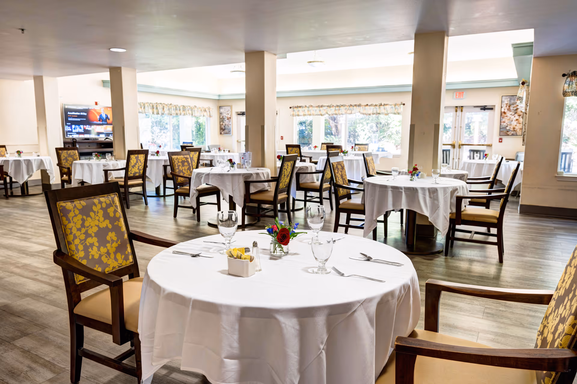 A bright dining room with round tables covered in white tablecloths, each set with glassware, silverware, and small floral centerpieces. The room has wooden chairs with yellow floral upholstery, large windows letting in natural light, and a television mounted on the wall in the background.