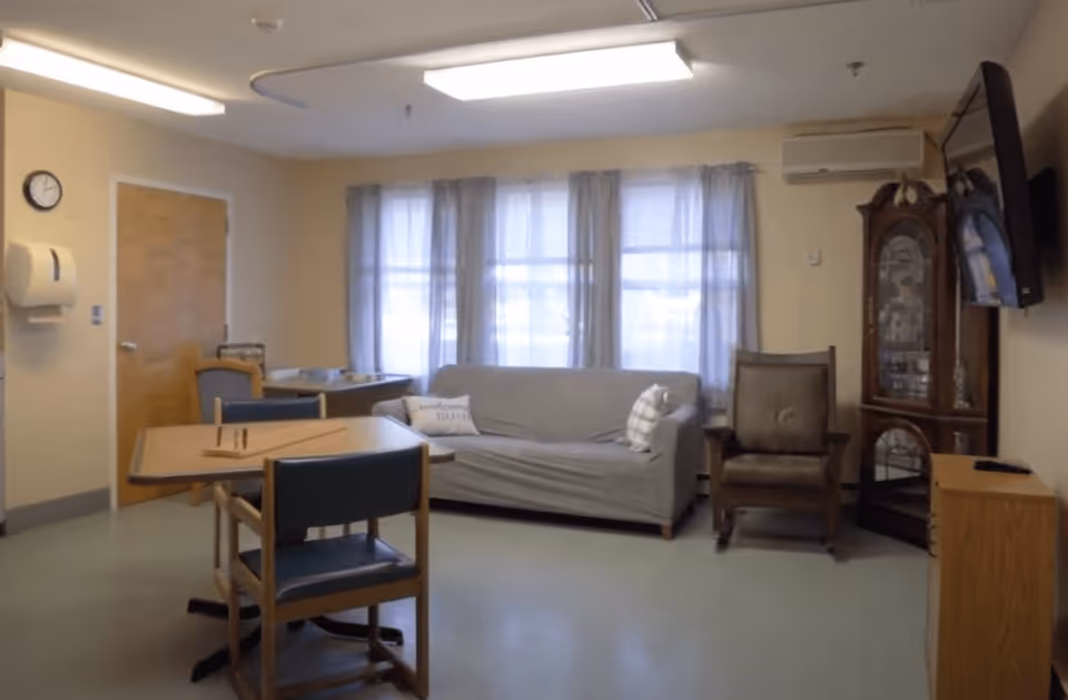 A cozy room with a gray sofa covered with a slipcover, two pillows, a wooden rocking chair, a wooden table with two chairs, a wall-mounted TV, a grandfather clock, and a window with sheer curtains letting in natural light.