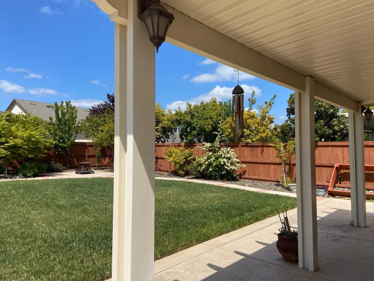 View of a backyard garden with a green lawn, various trees and shrubs along a wooden fence, and a covered patio with white support columns and hanging wind chimes under a blue sky with scattered clouds.