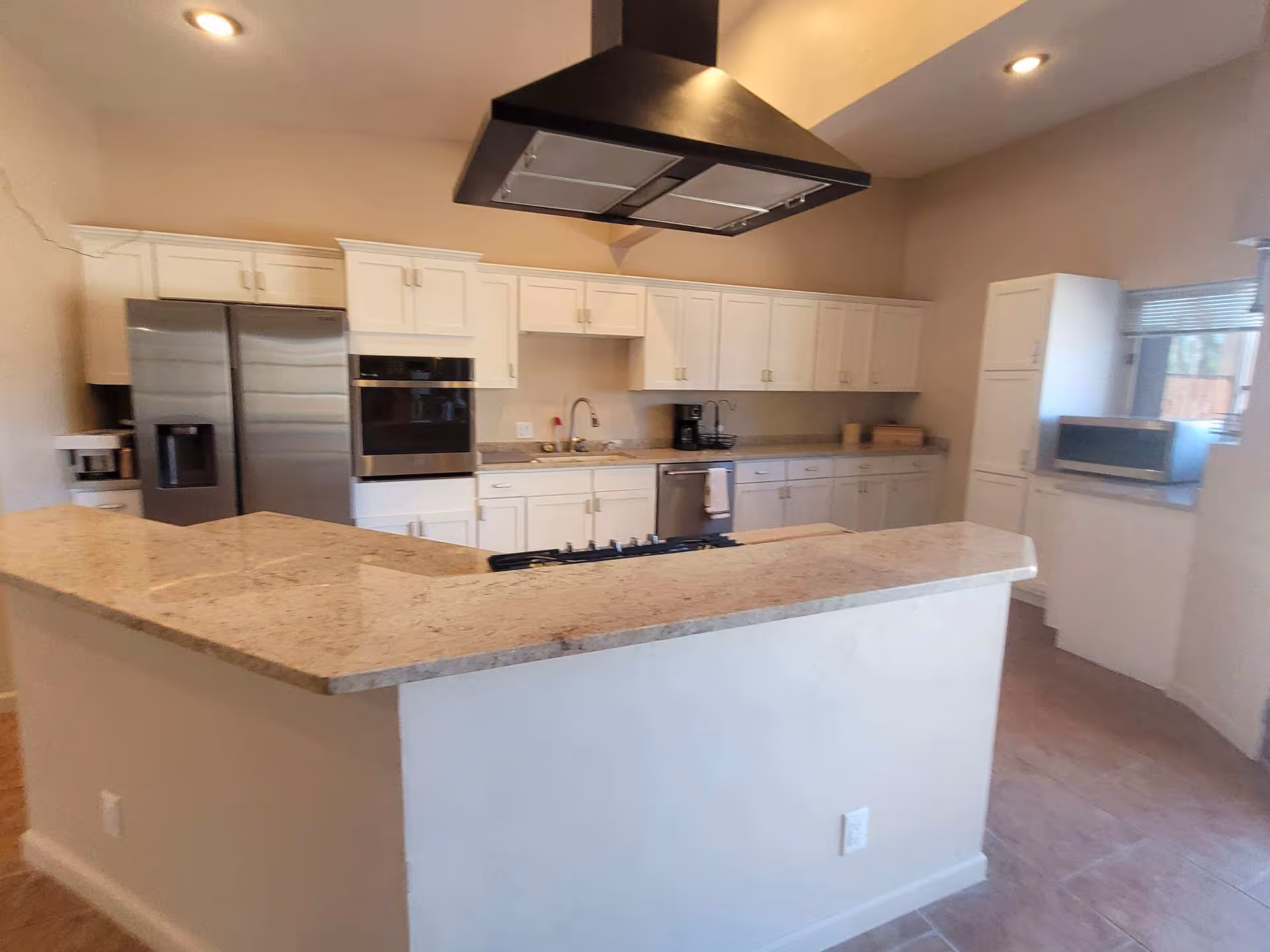Modern kitchen with white cabinets, a large granite countertop island with a built-in stove, stainless steel refrigerator, built-in oven, dishwasher, microwave, and a coffee maker. The kitchen has a beige tile floor and neutral-colored walls.
