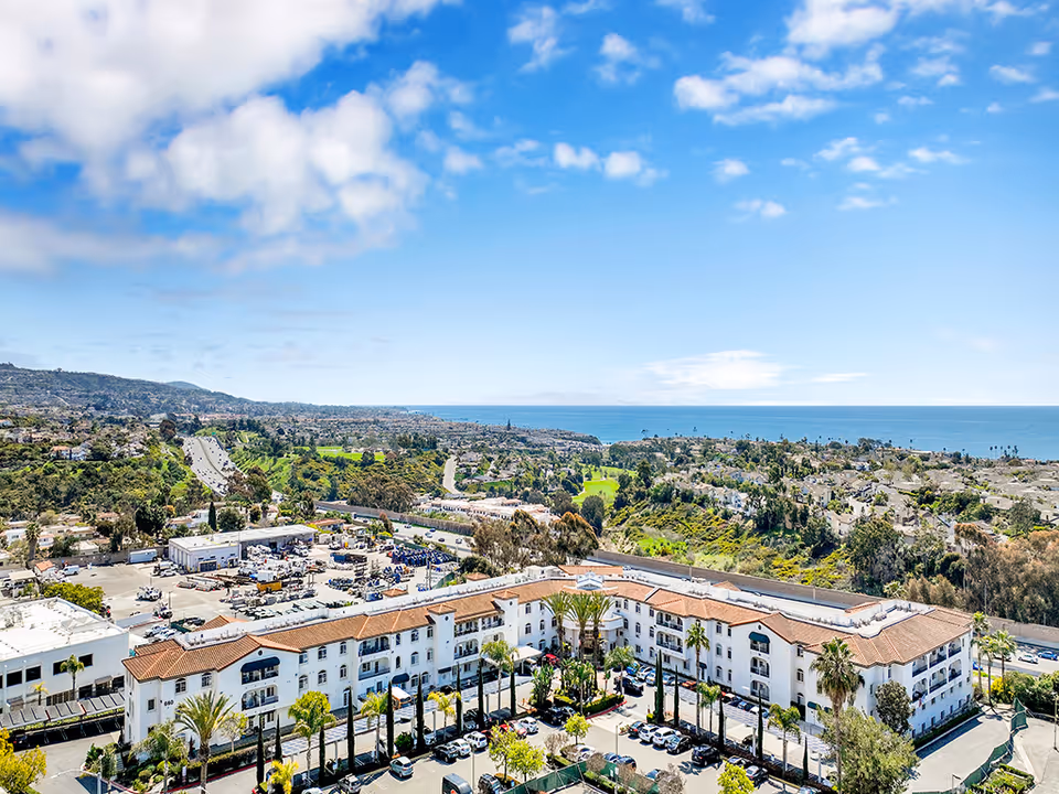 Aerial view of San Clemente Villas, a large white building with a red-tiled roof surrounded by palm trees and a parking lot. The background features a scenic landscape with green hills, residential areas, and the ocean under a partly cloudy blue sky.