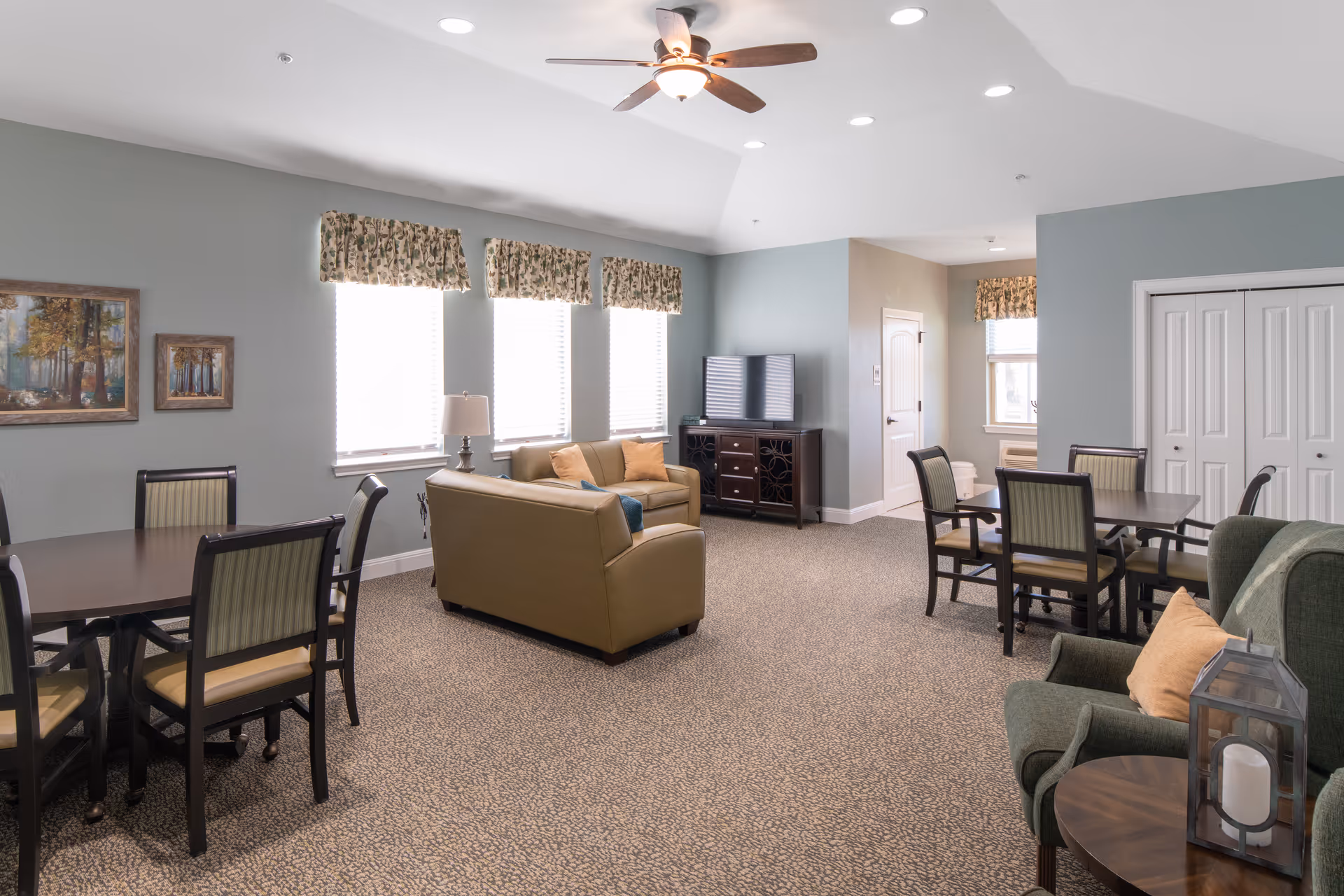 Bright communal living room with sofas, armchairs, dining tables and a TV in a senior living facility.