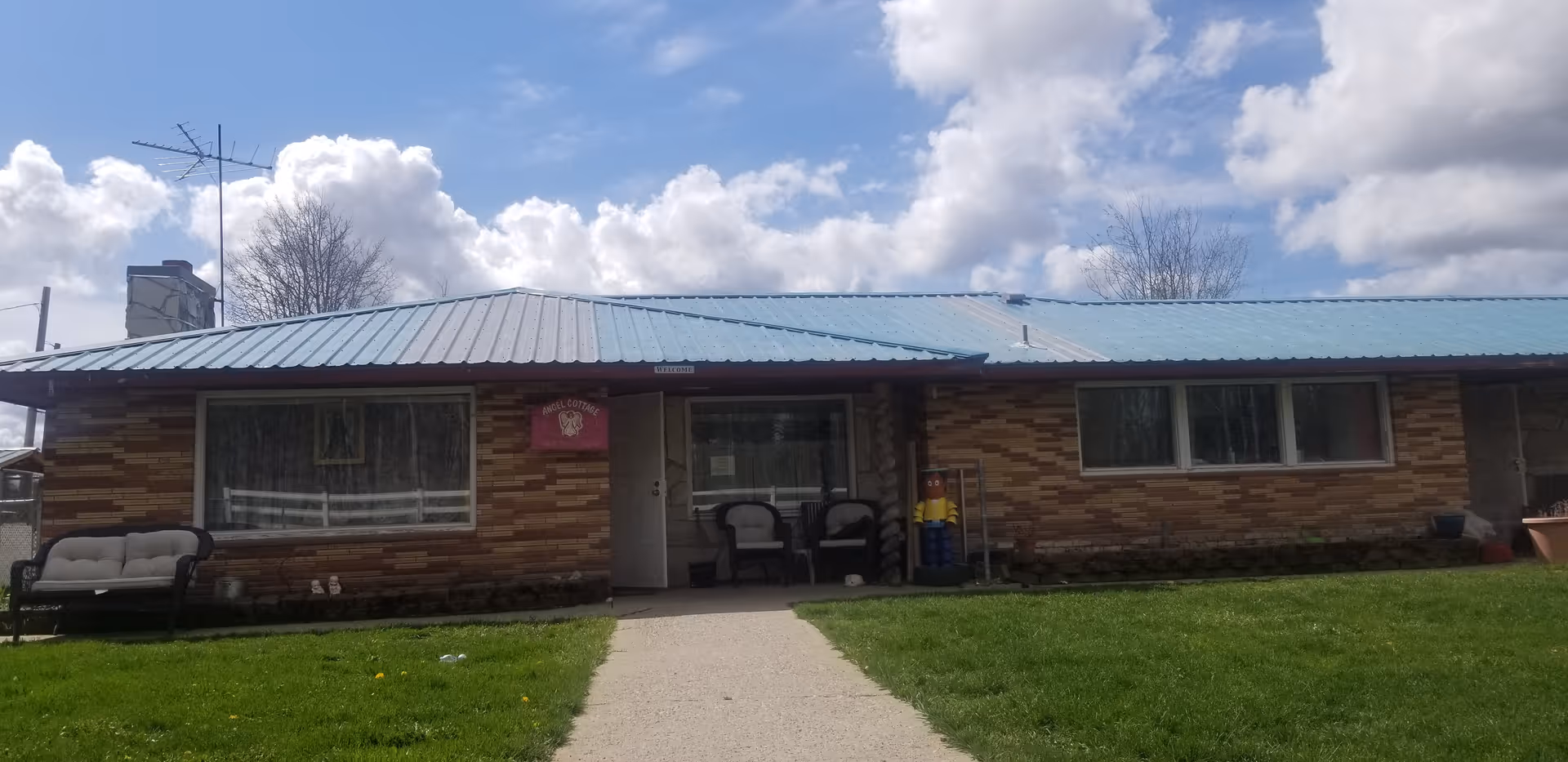 Single-story brick building with a light blue metal roof under a partly cloudy sky. There is a concrete walkway leading to the entrance, which has two chairs on the porch. A cushioned bench is placed on the left side of the building. A sign near the entrance reads 'Angel Cottage Adult Family Home'. The surrounding area has green grass and some bare trees in the background.