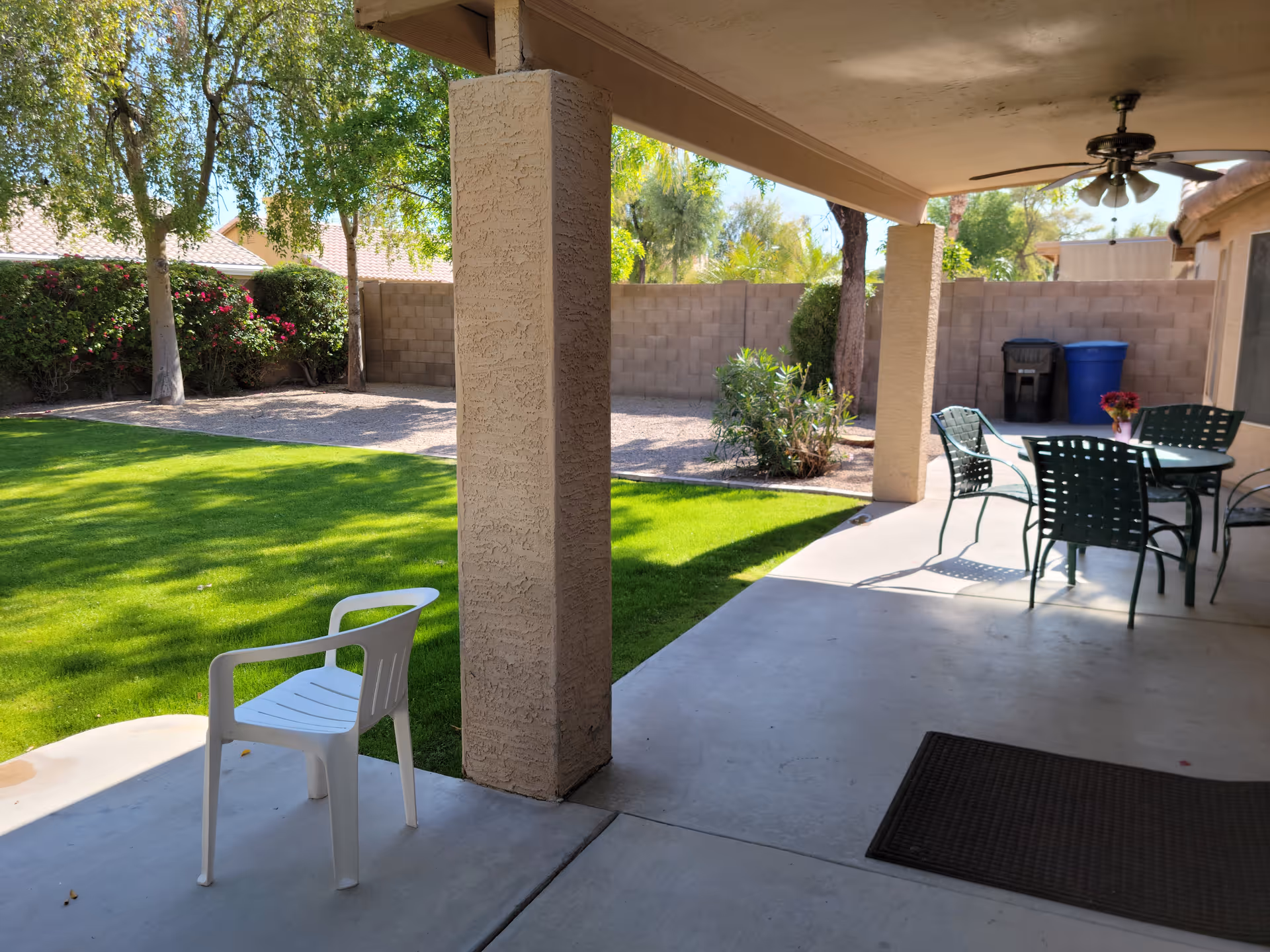 Covered patio area with a white plastic chair and a glass-top table with four green metal chairs. The patio overlooks a green lawn, trees, and bushes with a block wall fence in the background. There is a ceiling fan above the patio and two trash bins near the wall.
