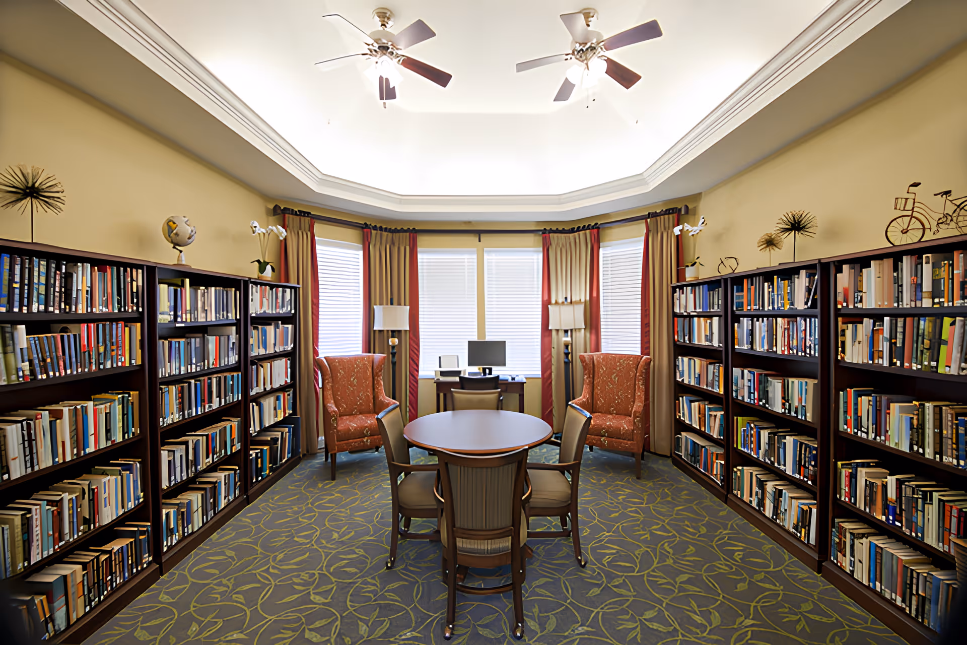 A cozy library room in a senior living facility with bookshelves filled with books lining both sides of the room. In the center, there is a round table with four chairs around it. At the far end, two upholstered armchairs are positioned near a window with closed blinds and curtains. A desk with a computer and two lamps is placed between the armchairs. The ceiling features two ceiling fans with lights.