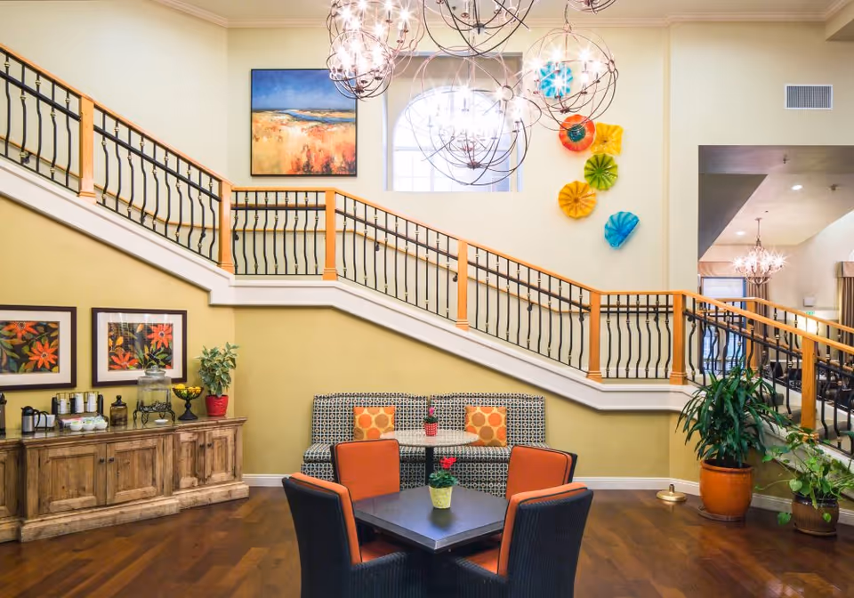 A bright and welcoming interior space featuring a staircase with wooden handrails and black balusters. Below the staircase is a seating area with a patterned bench and two orange cushioned chairs around a small table with a potted plant. The walls are decorated with colorful artwork, including framed floral prints and vibrant wall sculptures. There are also large, spherical chandeliers hanging from the ceiling, and several potted plants placed around the room.