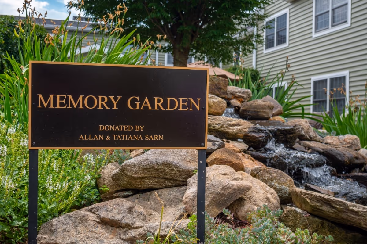 A black and gold sign reading 'Memory Garden Donated by Allan & Tatiana Sarn' is placed in front of a landscaped garden area with rocks, flowing water, green plants, and a tree. A building with beige siding and white-framed windows is visible in the background.
