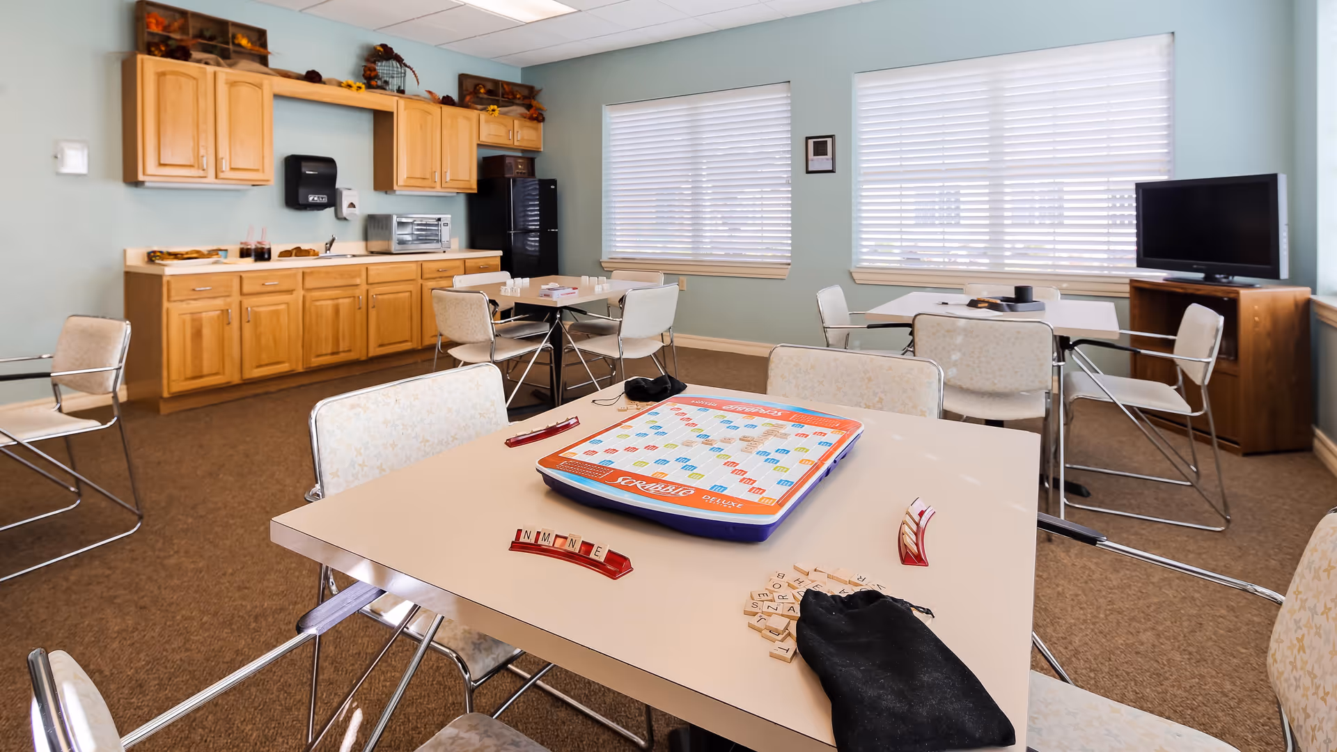 A bright room with light blue walls featuring several tables and chairs arranged for group activities. On the closest table, there is a Scrabble game set up with letter tiles and racks. The room has large windows with blinds, a TV on a wooden stand, and a kitchenette area with wooden cabinets, a microwave, and a refrigerator.