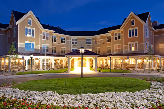 Exterior view of MorningStar Assisted Living & Memory Care at Jordan building at dusk, showing a well-lit three-story structure with multiple windows, a covered entrance, green lawn, and flower beds in the foreground.