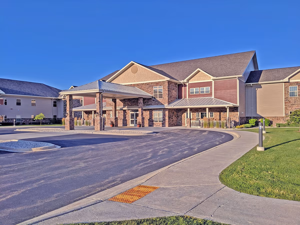Front exterior of a two-story brick and siding retirement community with a covered porte-cochere, curved driveway and sidewalk under a clear blue sky.