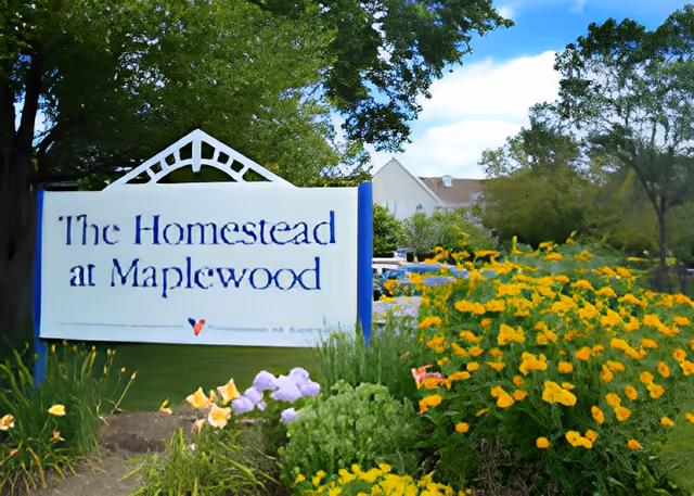 Entrance sign reading "The Homestead at Maplewood" surrounded by flowering plants and trees.