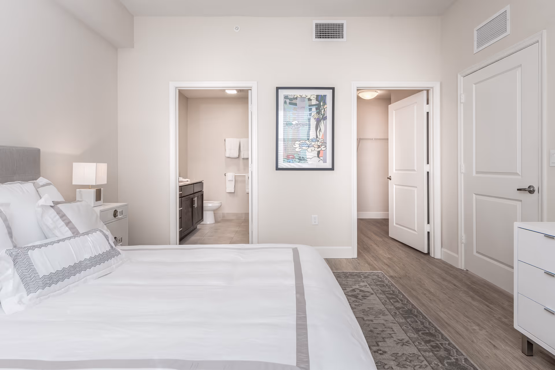 A bright and clean bedroom with a neatly made bed featuring white and gray bedding. To the left of the bed is a white nightstand with a modern lamp. The room has light-colored walls and wood flooring with a patterned rug. There are two open doorways on the far wall; the left doorway leads to a bathroom with a toilet, dark cabinetry, and towels hanging on the wall. The right doorway leads to a walk-in closet with an open door. A framed artwork hangs on the wall between the two doorways.