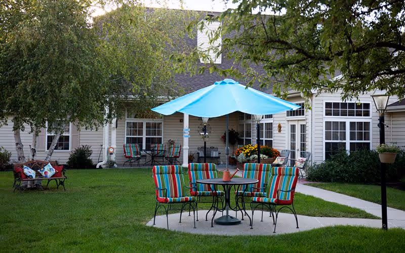 Outdoor seating area with a round table and four colorful striped cushioned chairs under a large blue umbrella on a concrete patio. In the background, there is a beige building with multiple windows and a covered porch with additional seating. Green grass and trees surround the area.