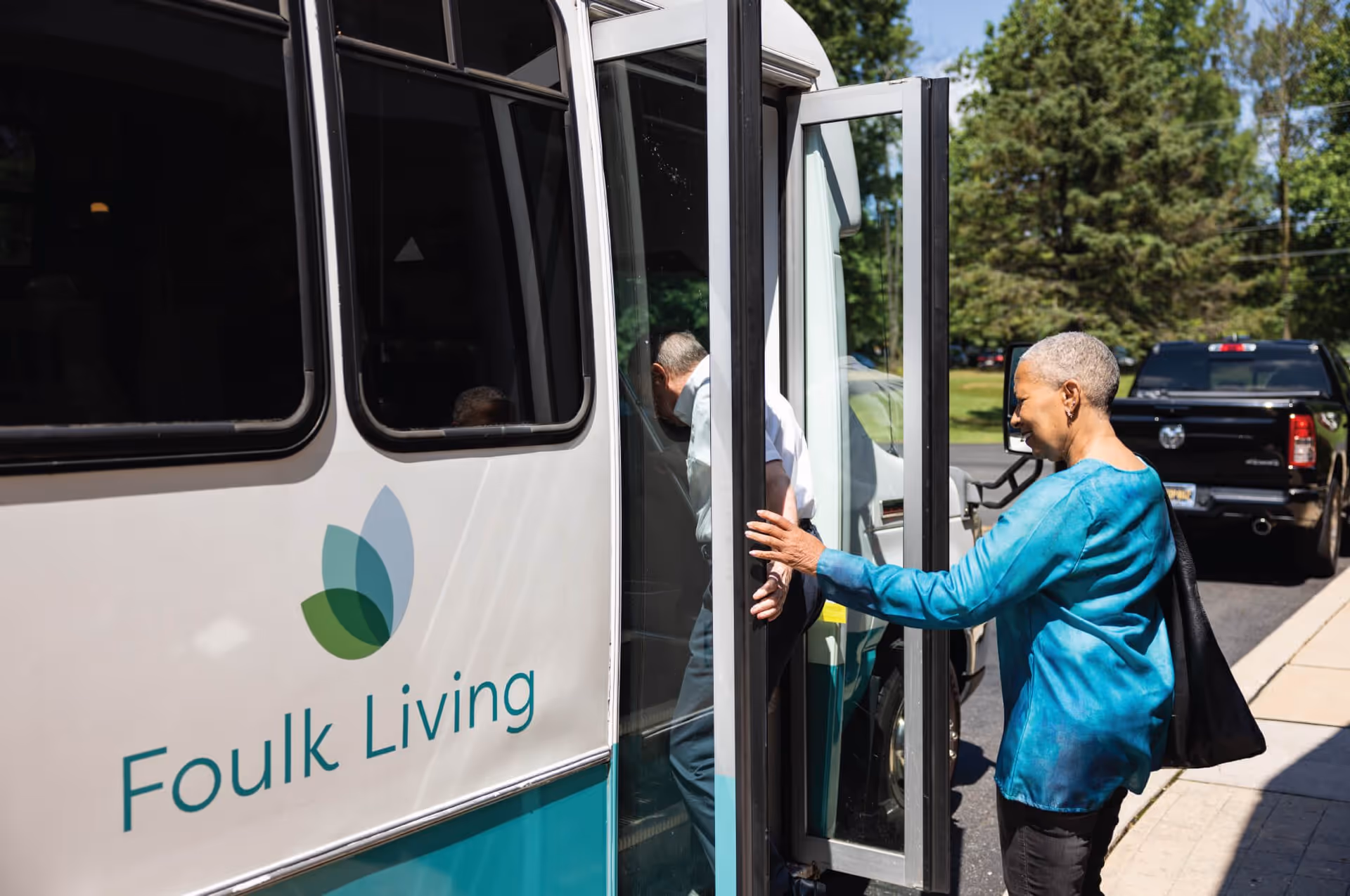 An elderly woman in a blue top holding the door open for an elderly man boarding a Foulk Living shuttle bus outside on a sunny day with trees and parked vehicles in the background.