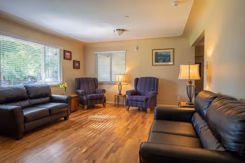 A cozy living room with two black leather sofas and two purple armchairs arranged around a wooden floor. There are two table lamps on side tables, framed pictures on the walls, and windows letting in natural light.