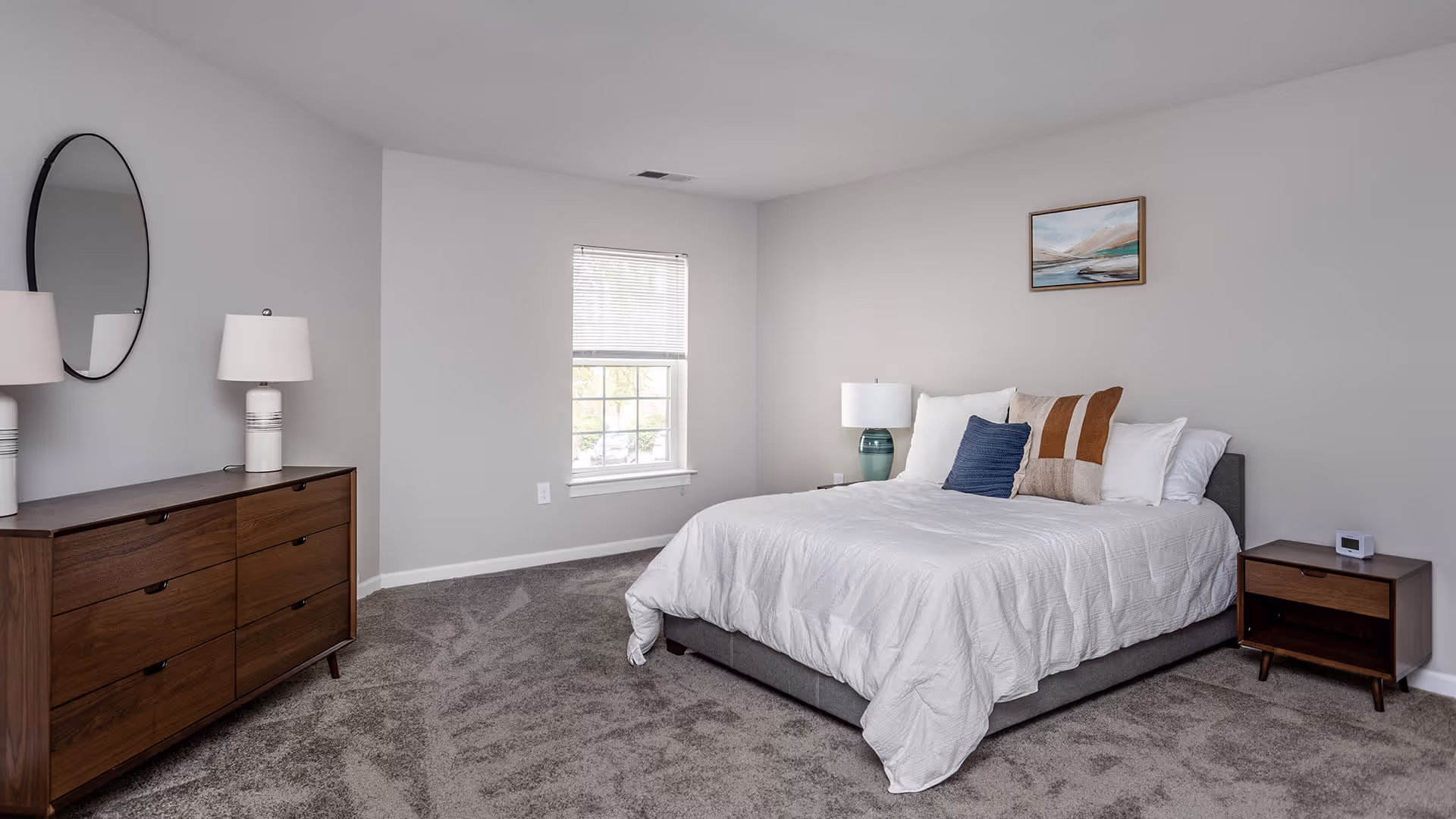 A neatly arranged bedroom with a large bed covered in white bedding and multiple pillows. There is a wooden nightstand with a lamp and a small clock on the right side of the bed. On the left side of the room, there is a wooden dresser with two lamps and a round mirror above it. A window with blinds is centered on the far wall, and a framed landscape painting hangs above the bed. The room has light gray walls and carpeted flooring.