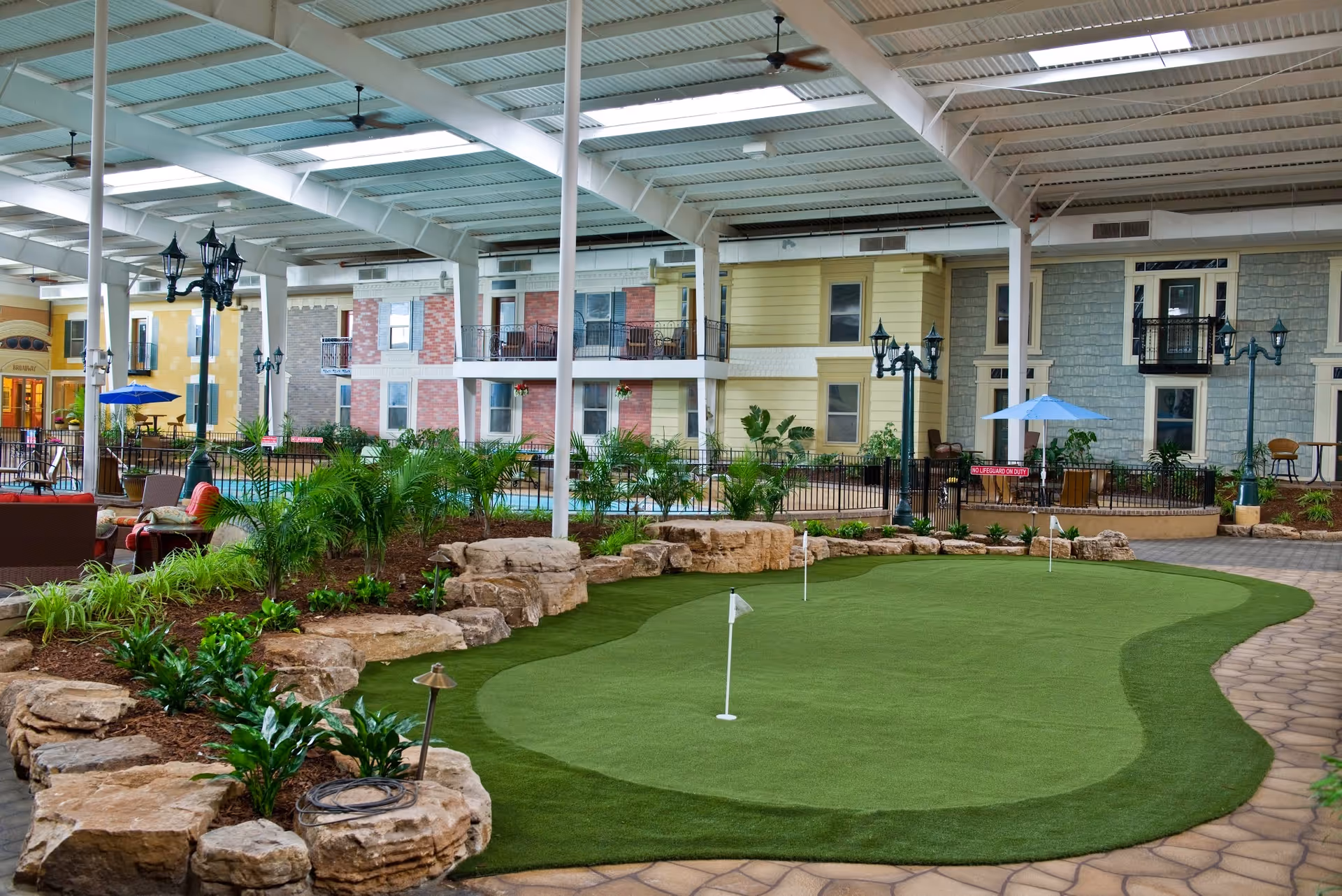 Indoor putting green surrounded by landscaping with rocks and plants, under a large covered roof with ceiling fans. In the background, there are multi-story residential-style buildings with balconies, outdoor seating areas, and streetlamp-style lights.