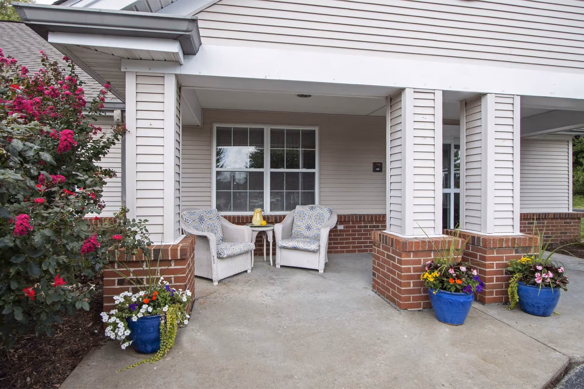Covered outdoor seating area with two cushioned wicker chairs and a small round table holding a pitcher and glass of lemonade. The area is framed by brick pillars and beige siding. There are blue flower pots with colorful flowers on the concrete floor and a bush with bright pink flowers to the left.