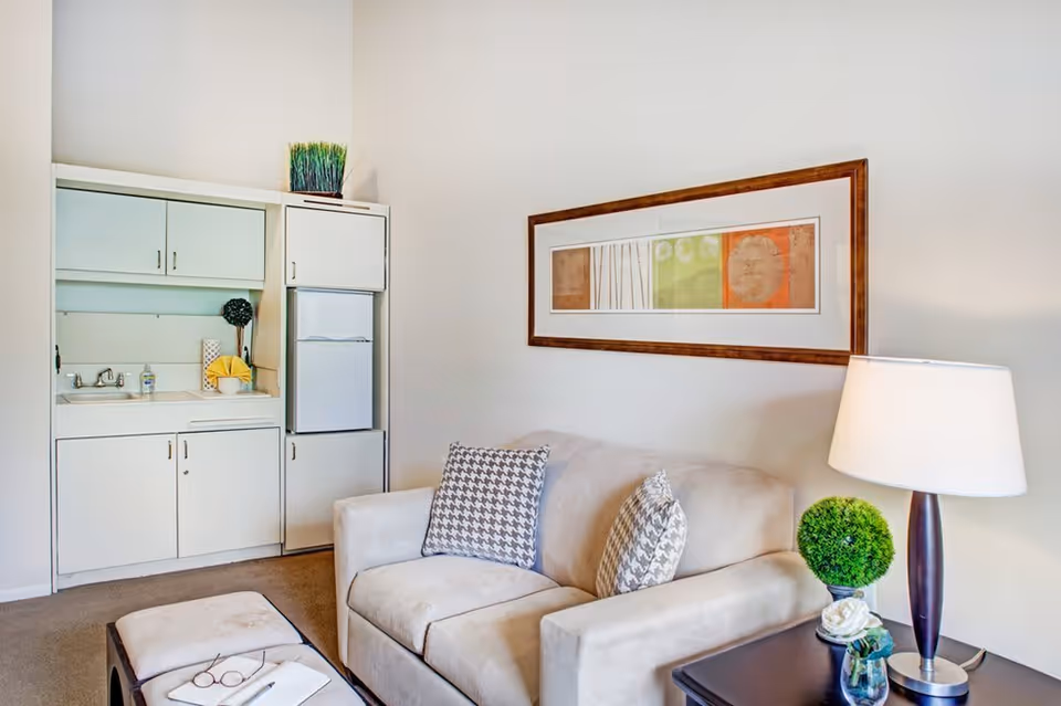 A cozy living room area with a beige sofa adorned with two patterned pillows, a dark wooden side table with a lamp and a small green plant, and a small kitchenette with white cabinets, a sink, and a refrigerator. A framed abstract artwork hangs on the wall above the sofa.