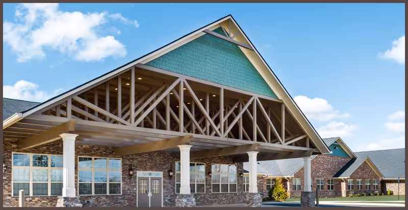 Exterior view of a senior living facility named Antebellum James Burgess showing a large covered entrance with white columns, stone walls, multiple windows, and a blue-green gabled roof under a partly cloudy sky.