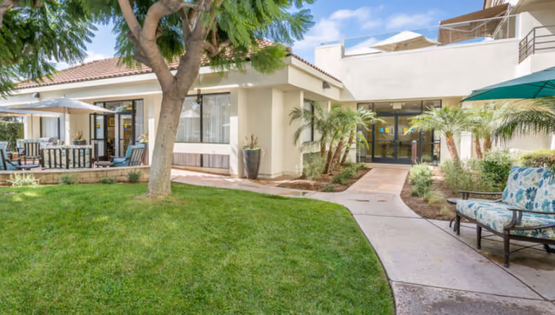 Outdoor courtyard with lawn, patio seating and a walkway leading to the entrance of an assisted living building.