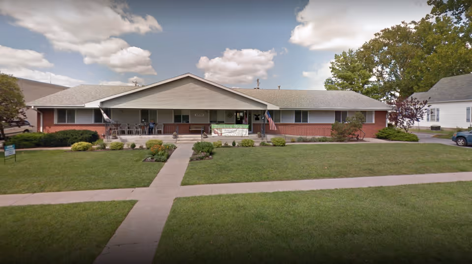 Single-story brick and siding building with a covered front porch, several chairs on the porch, an American flag, and a green lawn with a concrete walkway leading to the entrance under a partly cloudy sky.