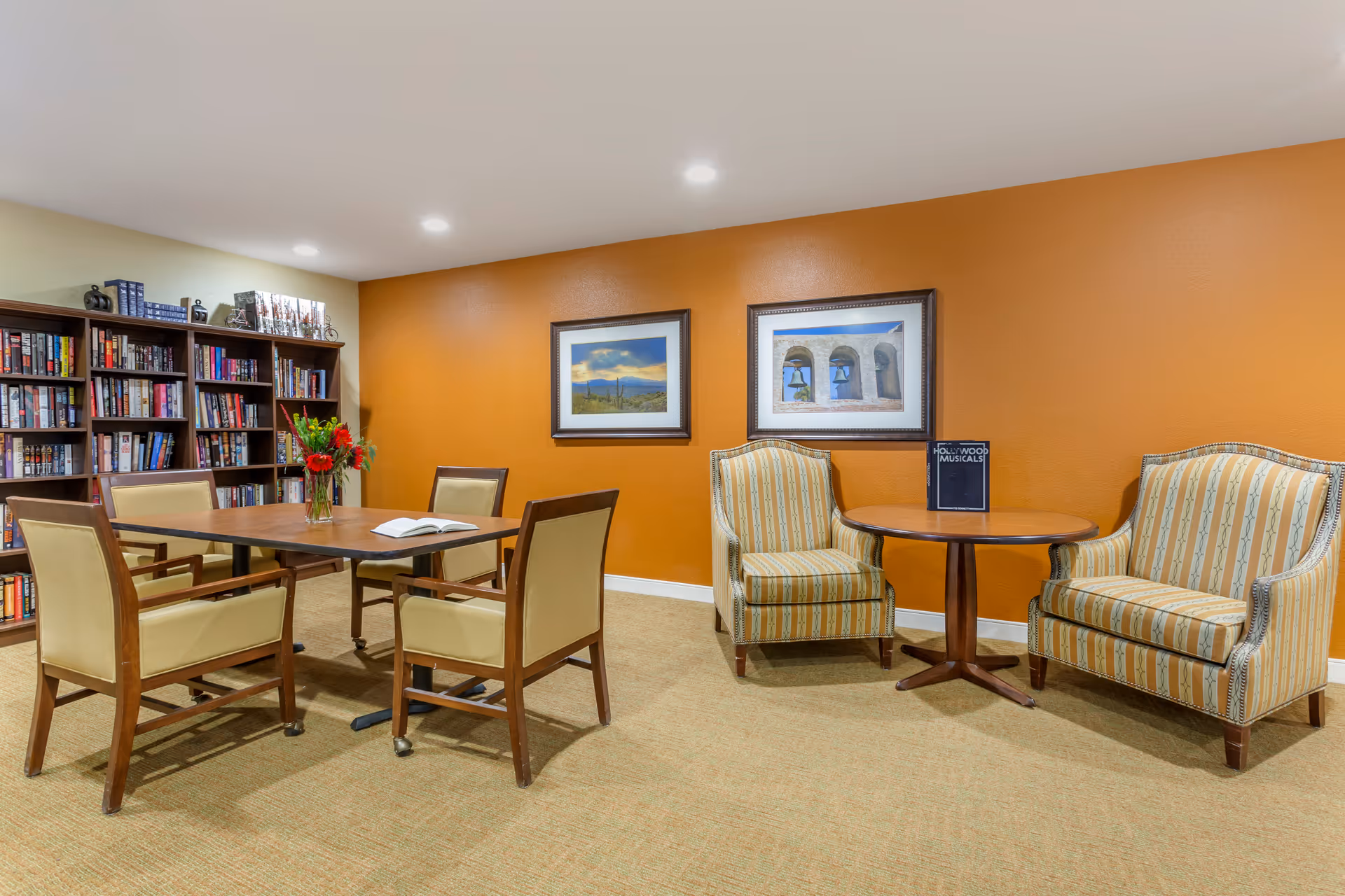 A cozy reading and sitting area with a wooden table surrounded by four beige cushioned chairs on the left, and two striped armchairs with a small round wooden table between them on the right. Behind the table on the left is a bookshelf filled with books and decorative items. The walls are painted orange and light yellow, and two framed pictures hang on the orange wall.