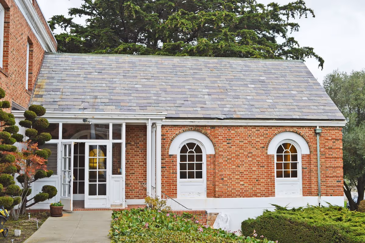 Exterior view of a brick building with a slate roof, featuring two arched windows with white trim and a glass door entrance. There are manicured bushes and a paved walkway leading to the entrance, with trees visible in the background.