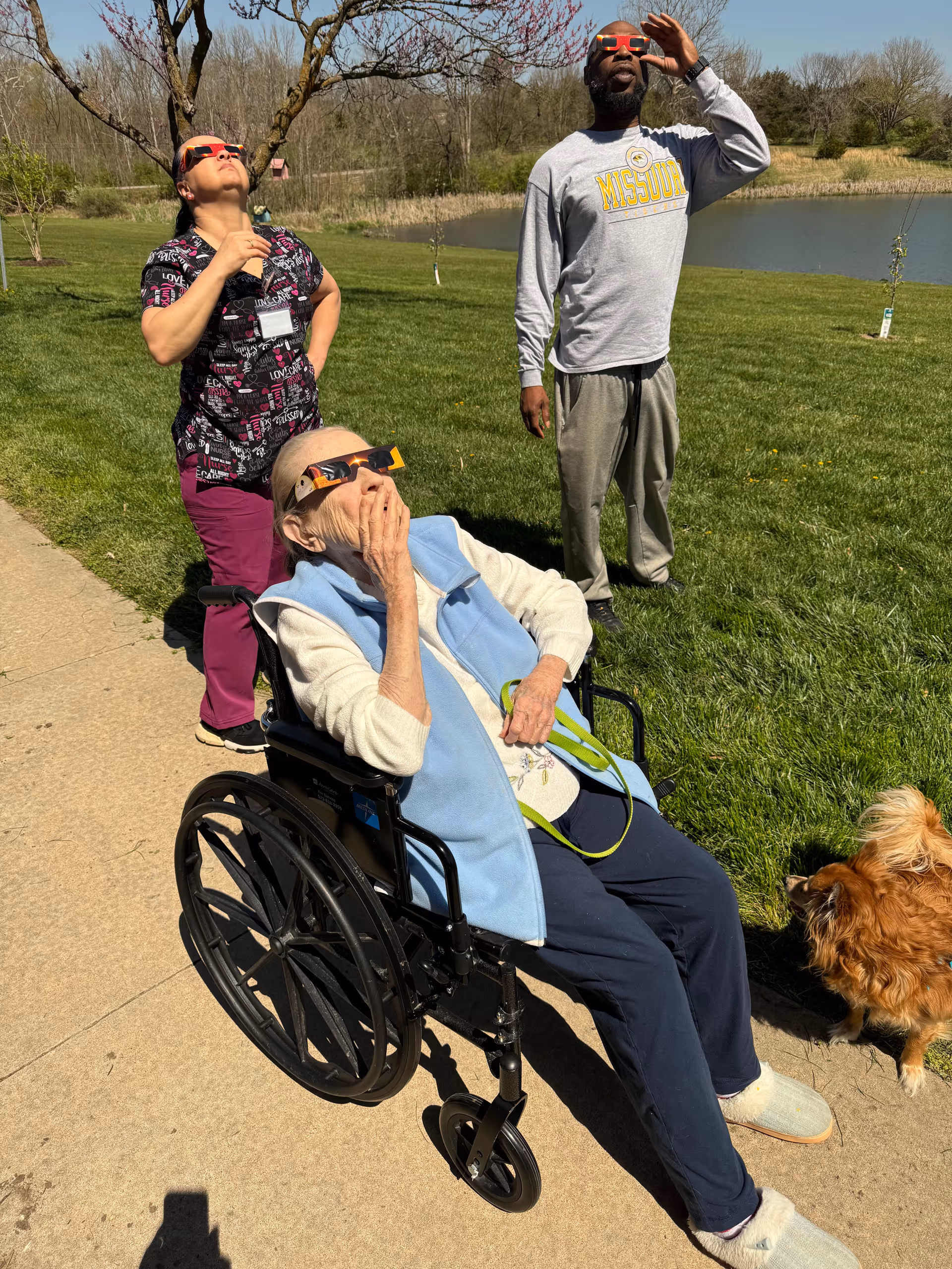 Three people outdoors on a sunny day near a pond, all wearing solar eclipse glasses and looking up at the sky. One elderly person is seated in a wheelchair wearing a light blue vest, while a woman in scrubs and a man in casual clothing stand behind her. A small brown dog is also present on the grass nearby.