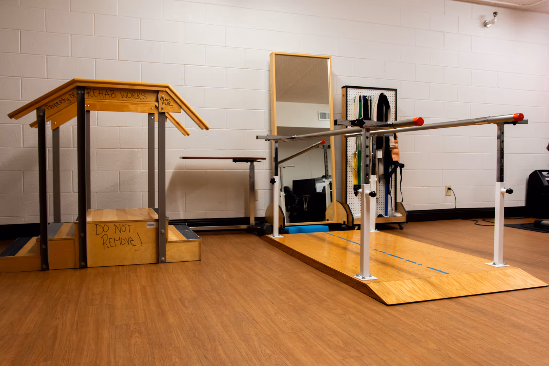 A rehabilitation room with wooden parallel bars on a platform for walking exercises, a wooden step structure labeled 'REHAB WORKS' and 'DO NOT REMOVE', a large mirror, and a pegboard holding various therapy equipment against a white brick wall.