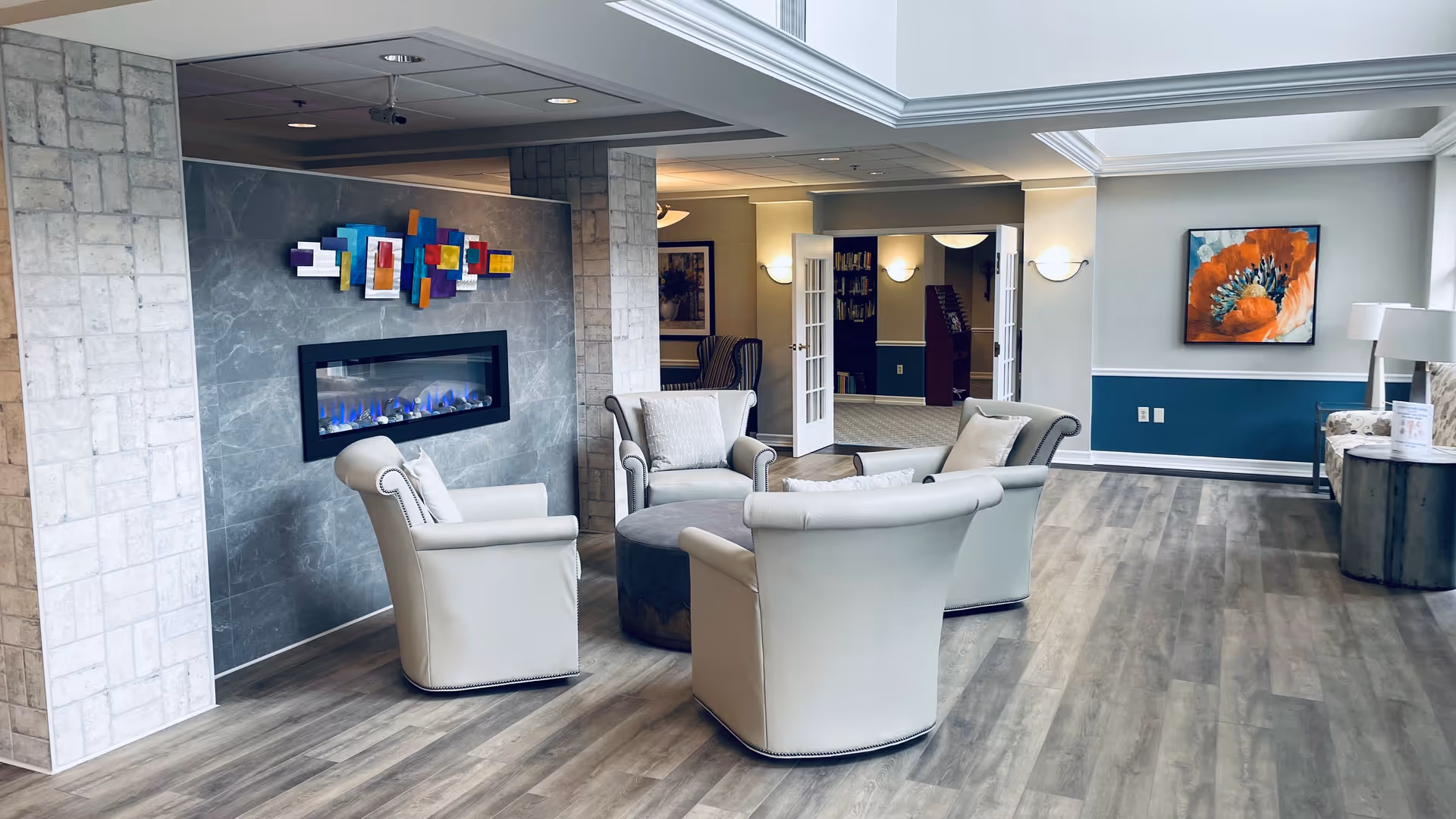 A modern and cozy seating area in a senior living facility with four beige armchairs arranged around a round ottoman in front of a wall-mounted electric fireplace. Above the fireplace is a colorful abstract wall art. The room has wood flooring, light gray walls with white trim, and a large window letting in natural light. In the background, there is an open doorway leading to another room with bookshelves and additional seating. A large floral painting and a floor lamp are visible on the right side.