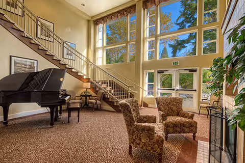 Sunlit communal lounge with a grand piano, patterned armchairs, a staircase, and large floor-to-ceiling windows.