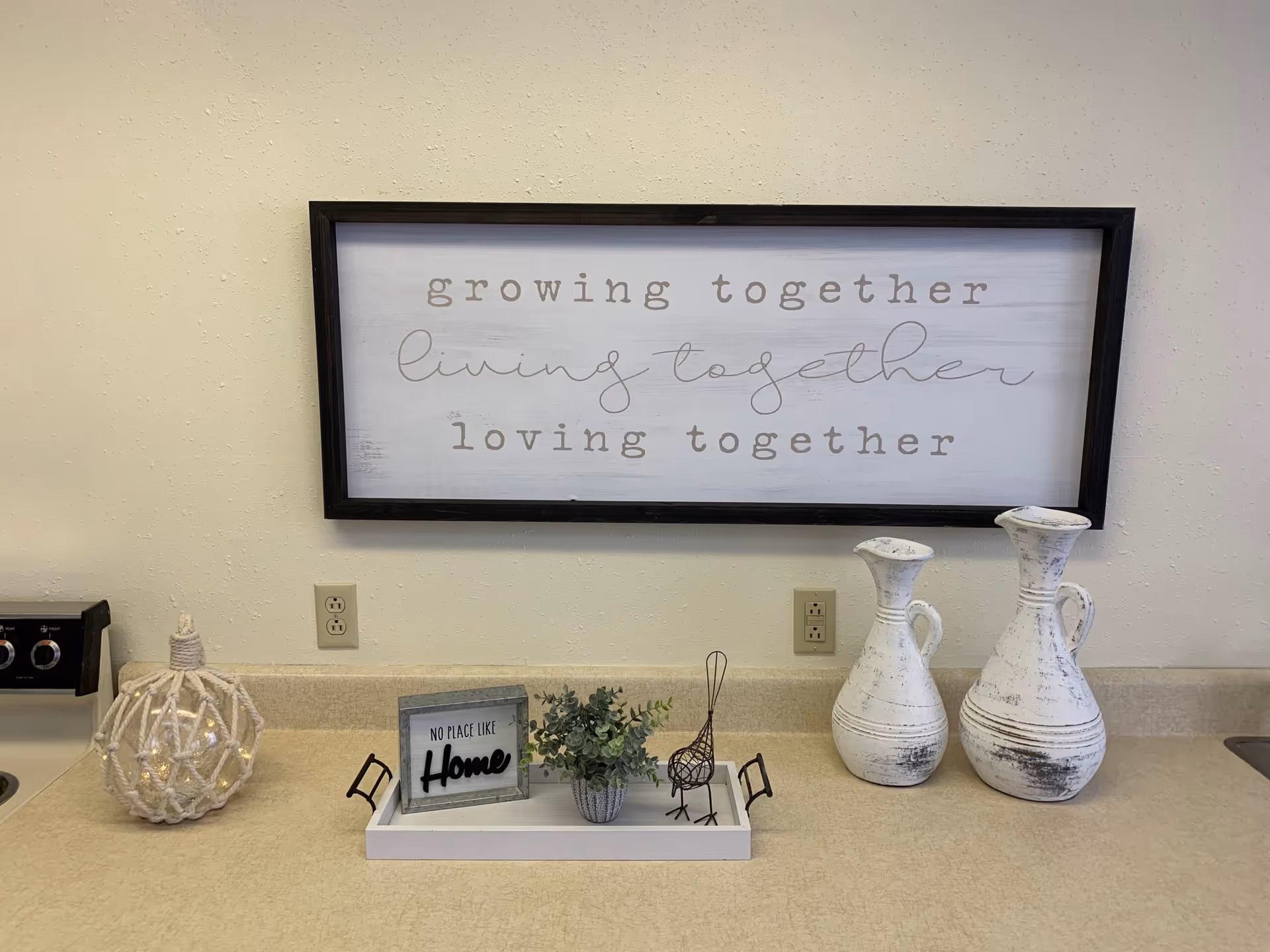 Decorated kitchen countertop with a large framed sign on the wall, two white distressed vases, a tray with a small plant and 'No place like Home' sign, and other tabletop decor.