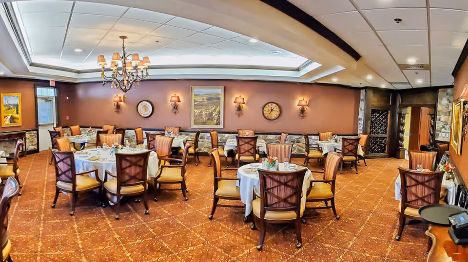 Formal dining room with round tables set with white tablecloths, upholstered wooden chairs, a chandelier, and warm wall decor.