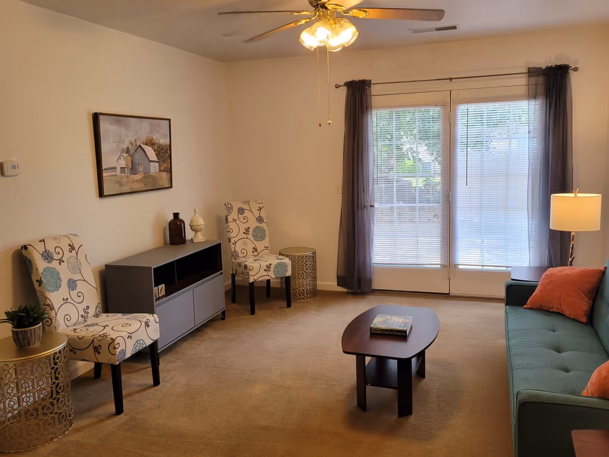 A living room with patterned accent chairs, a gray media console, a teal sofa with orange pillows, a coffee table, and glass doors with blinds.