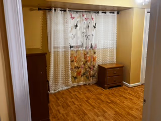 Empty small bedroom with hardwood floors, yellow walls, a window with floral sheer curtains, a dresser and a nightstand.