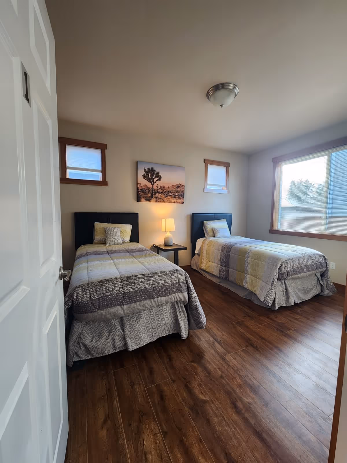 A bedroom with two single beds, each with a patterned comforter in shades of gray, yellow, and white. Between the beds is a small black nightstand with a lamp. Above the nightstand is a wall picture of a tree in a desert landscape. The room has wooden flooring, two small windows with blinds on the back wall, and a large window on the right side letting in natural light. The door to the room is partially open.