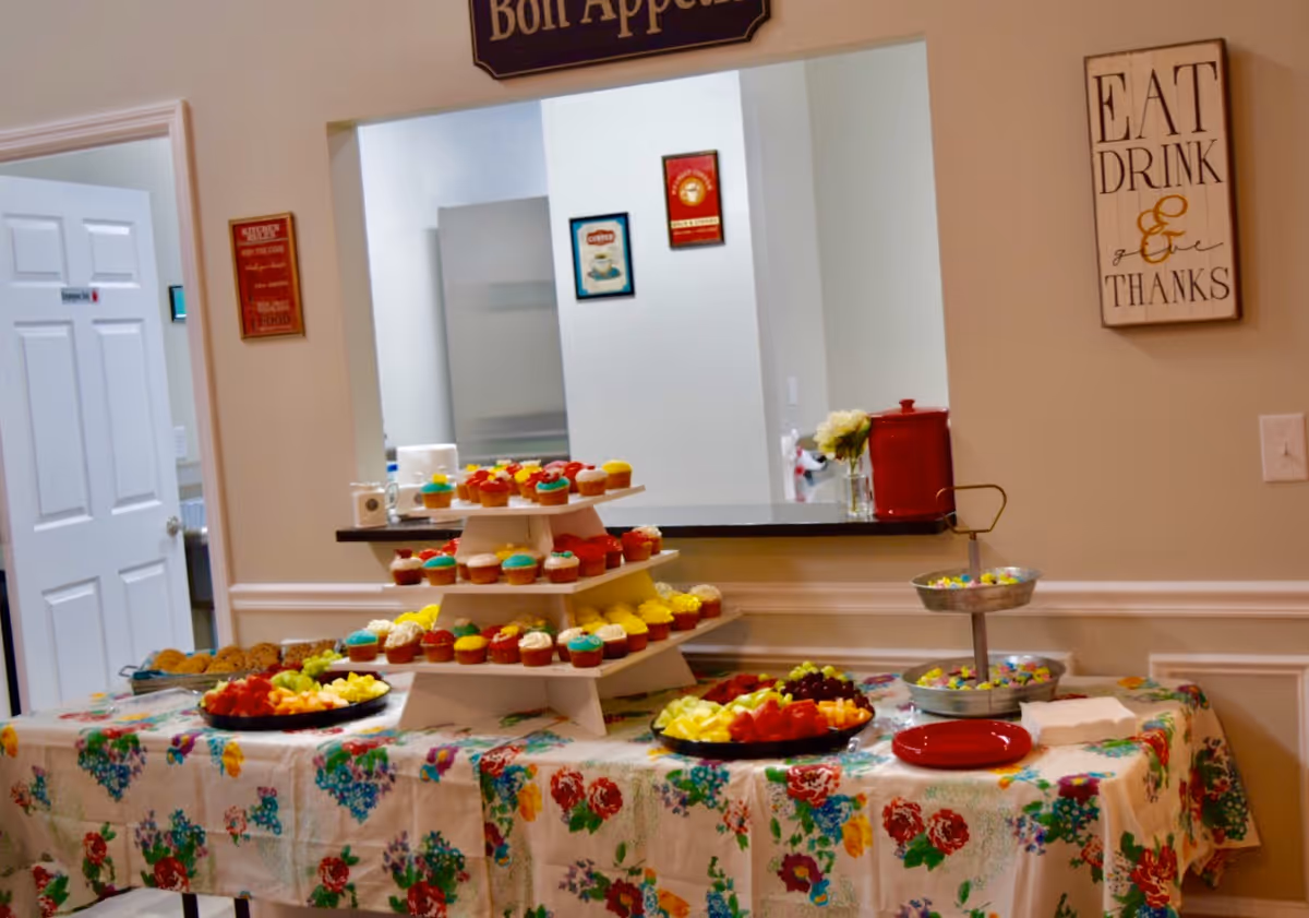 Decorated buffet table with a tiered display of colorful cupcakes, fruit platters, and treats in a communal dining area with wall signs.