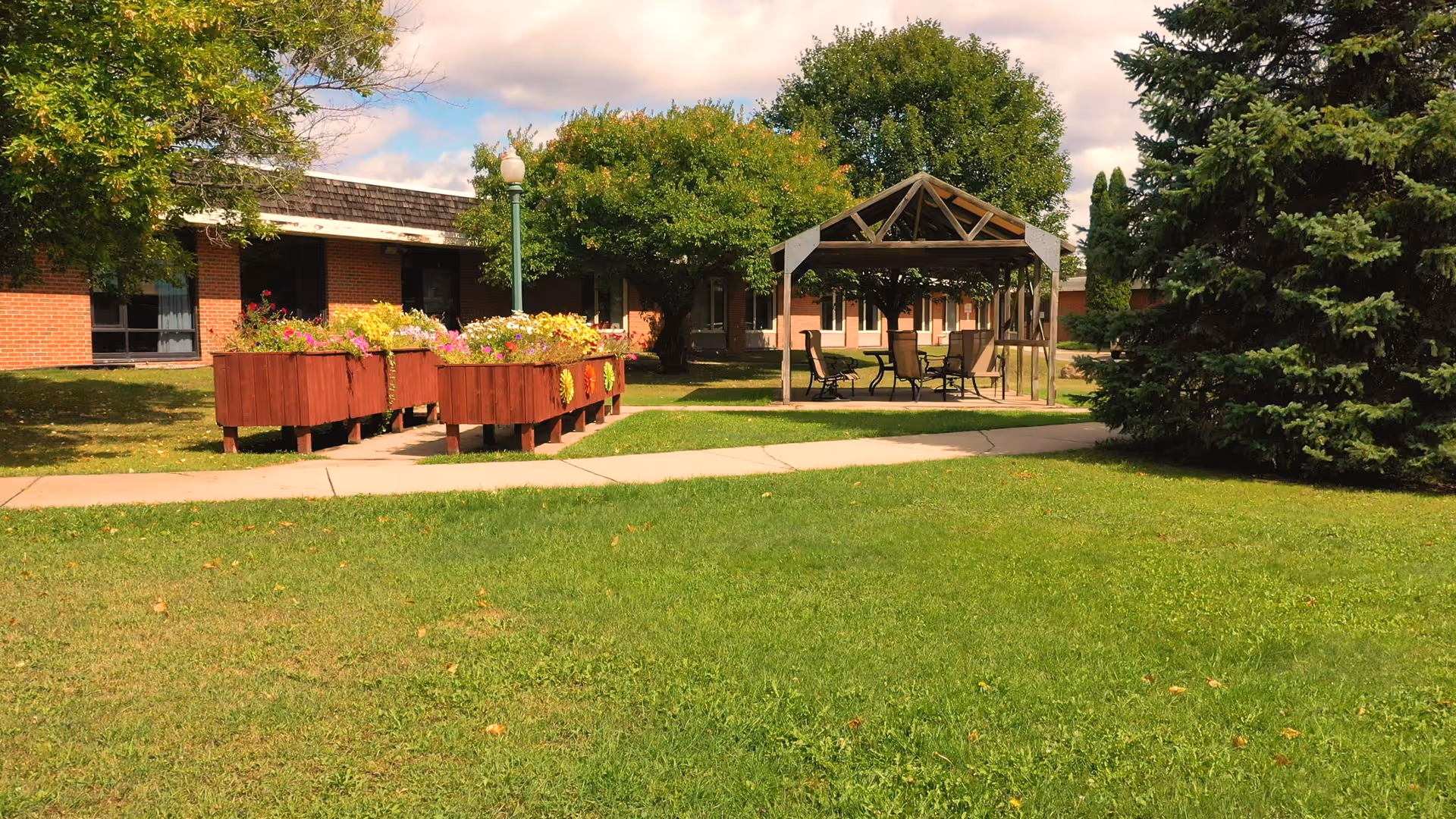 Outdoor garden area at The Waterview Pines featuring raised flower beds with colorful flowers, a paved walkway, a wooden gazebo with a table and chairs underneath, surrounded by green grass, trees, and a brick building in the background.