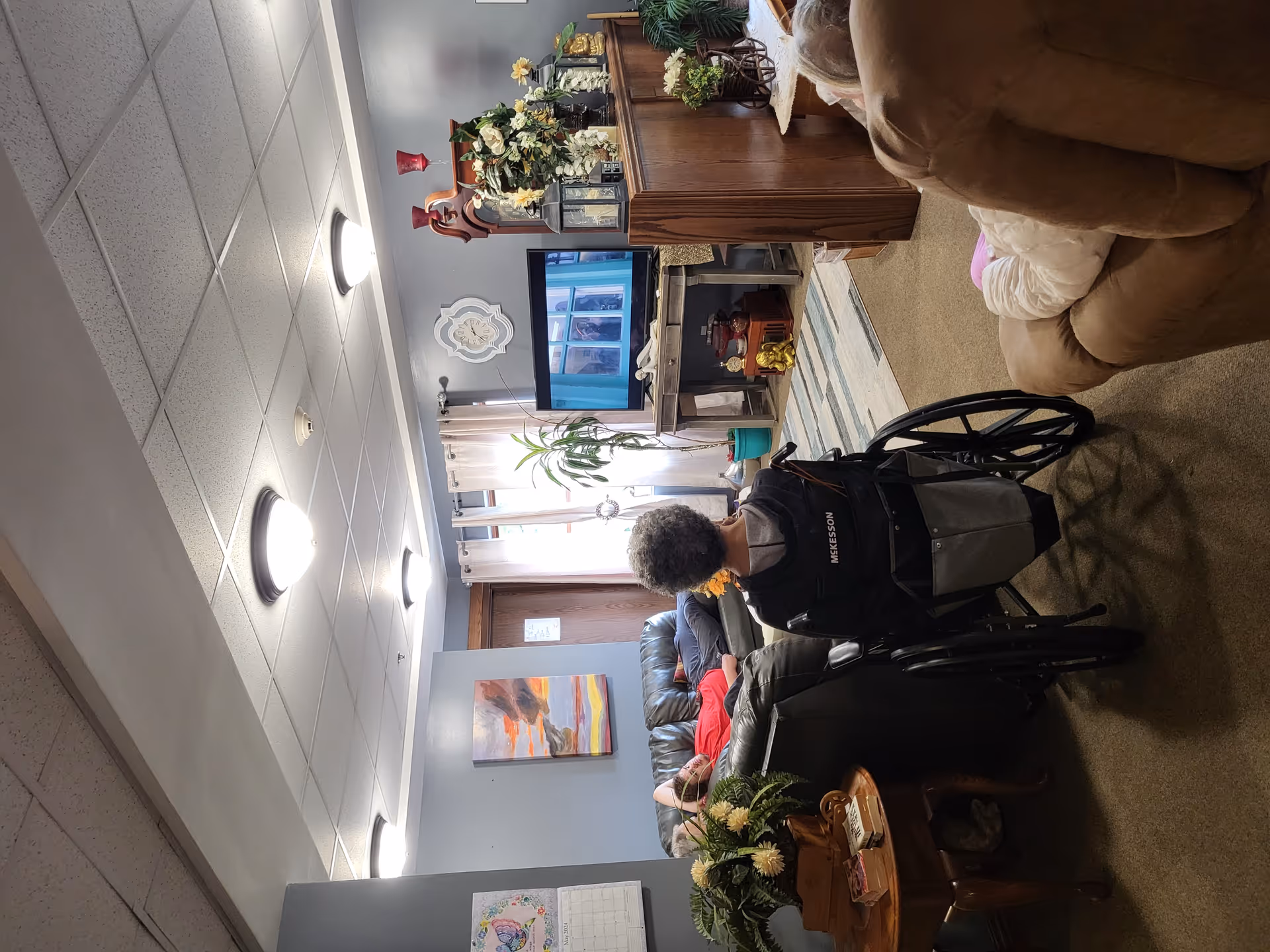 A cozy living room area in a senior living facility with a person in a wheelchair facing a television, another person reclining on a leather couch, and a third person seated in a recliner. The room has soft lighting, a wall clock, curtains on the window, decorative plants, and various ornaments on a wooden divider.