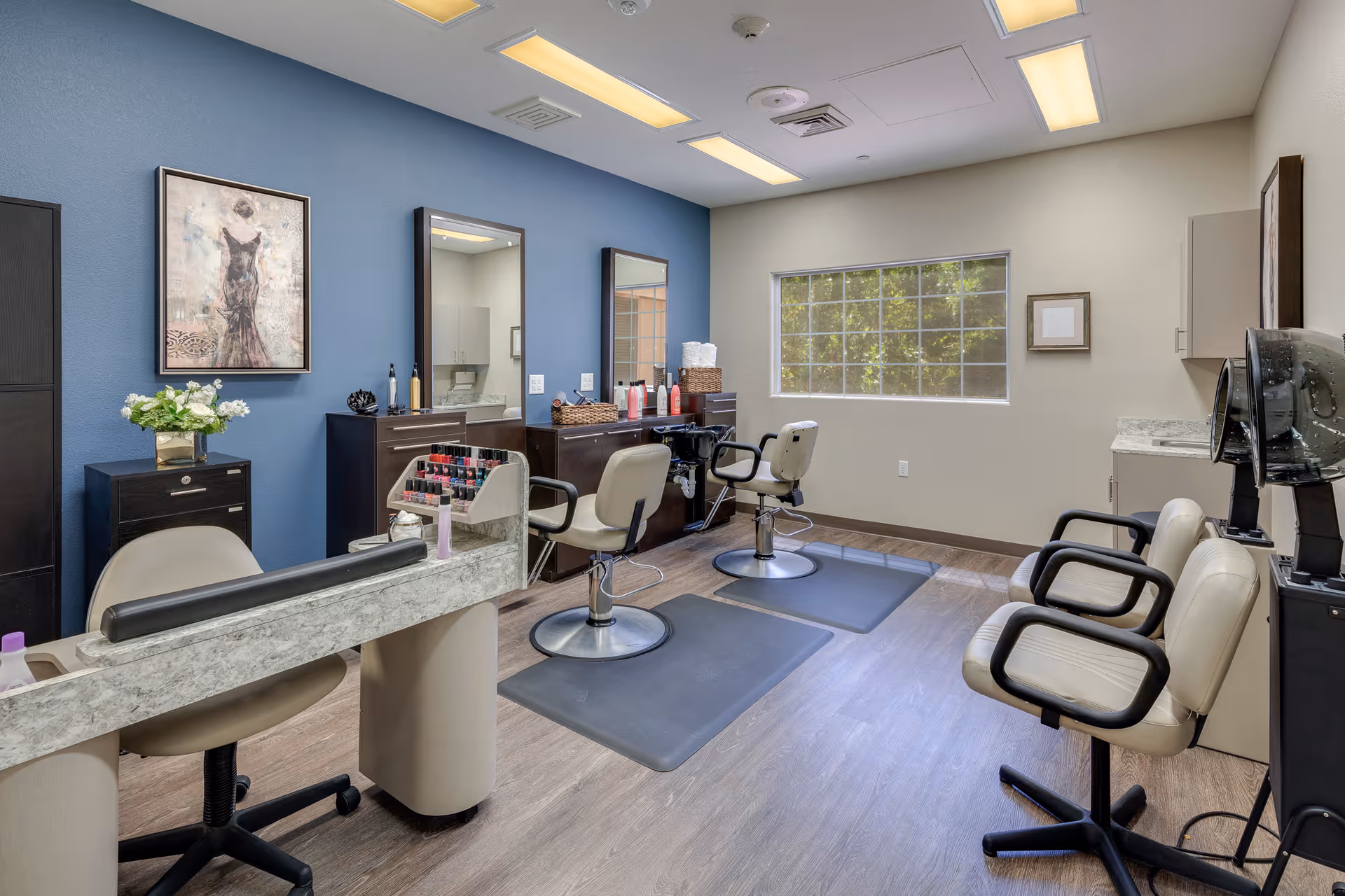 Interior of a salon room with two styling chairs in front of mirrors, a manicure station with a chair, nail polish display, and two hair dryer chairs along the right wall. The room has wood flooring, a blue accent wall with framed artwork, and a window letting in natural light.