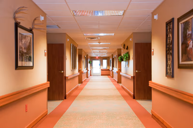 A long, well-lit hallway in a senior living facility with beige walls, wooden doors on both sides, handrails along the walls, framed artwork, and decorative plants. The hallway leads to a glass exit door at the far end.