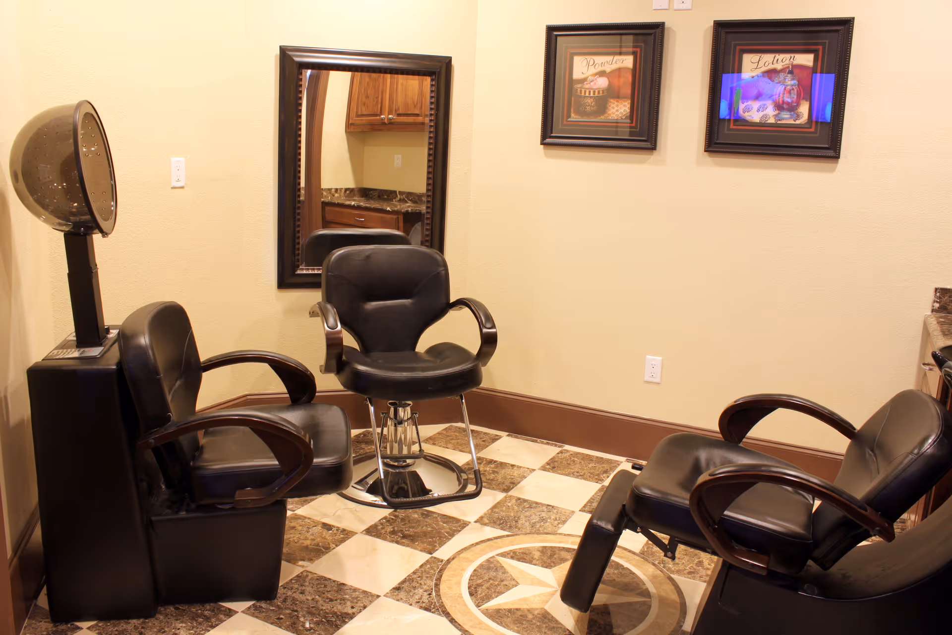 Interior view of a salon area with three black salon chairs, a hair dryer, a large mirror on the wall, two framed pictures labeled 'Powder' and 'Lotion', and a checkered floor with a star design.