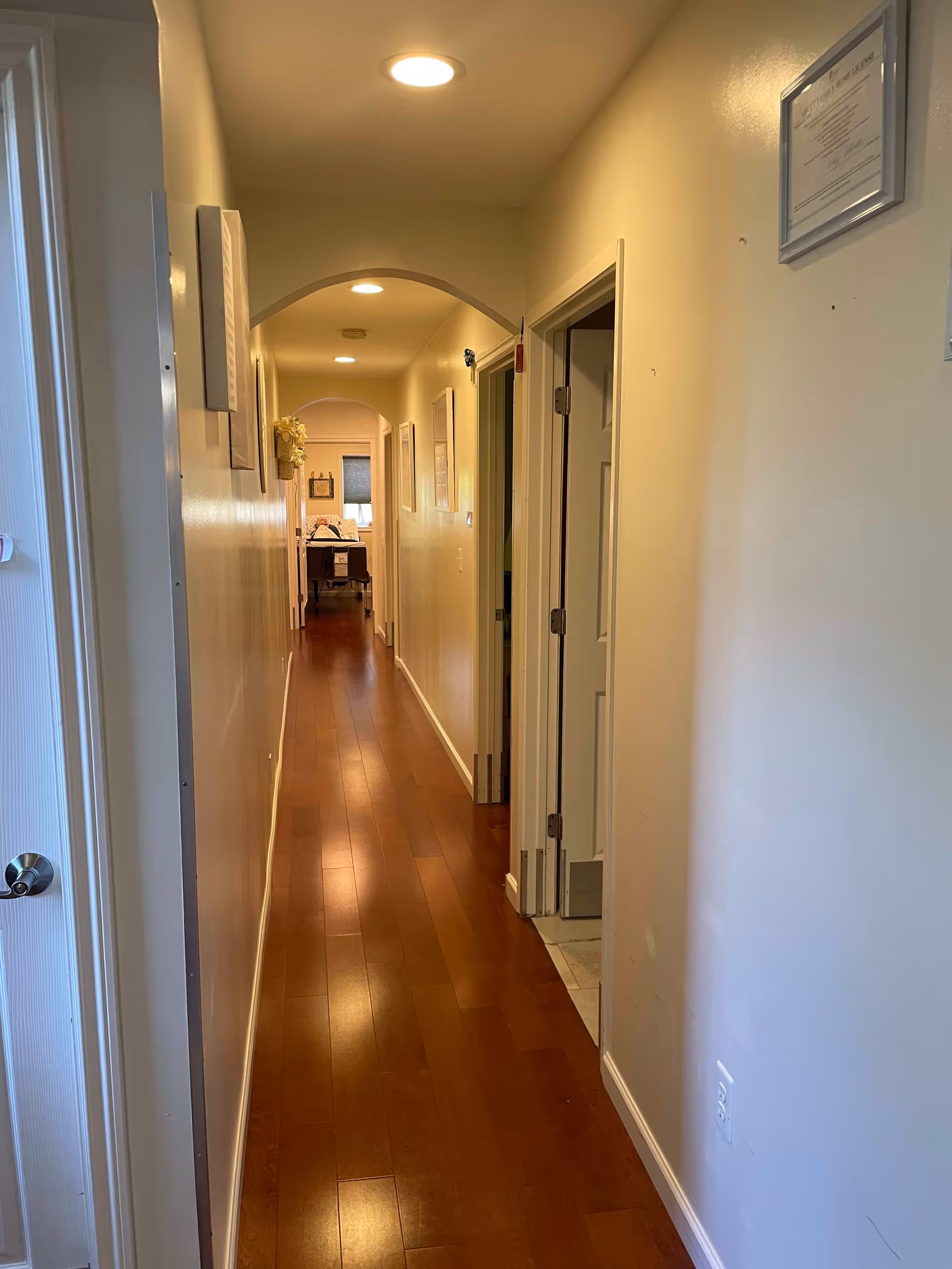 A long, narrow hallway with wooden flooring and cream-colored walls. The hallway is lit by ceiling lights and has several doorways on the right side. At the end of the hallway, there is a bed with white bedding visible in a room. Some framed pictures and a wall-mounted flower decoration are on the walls.