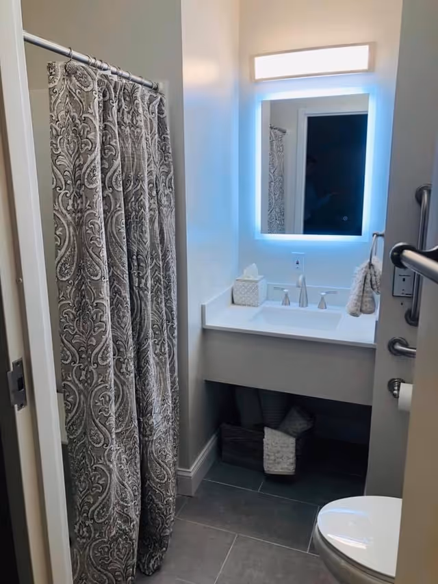 A small bathroom featuring a patterned shower curtain, a white sink with a modern faucet, a backlit rectangular mirror above the sink, a tissue box on the counter, a towel hanging on a grab bar, and a toilet partially visible in the foreground. There is a basket with rolled towels under the sink.