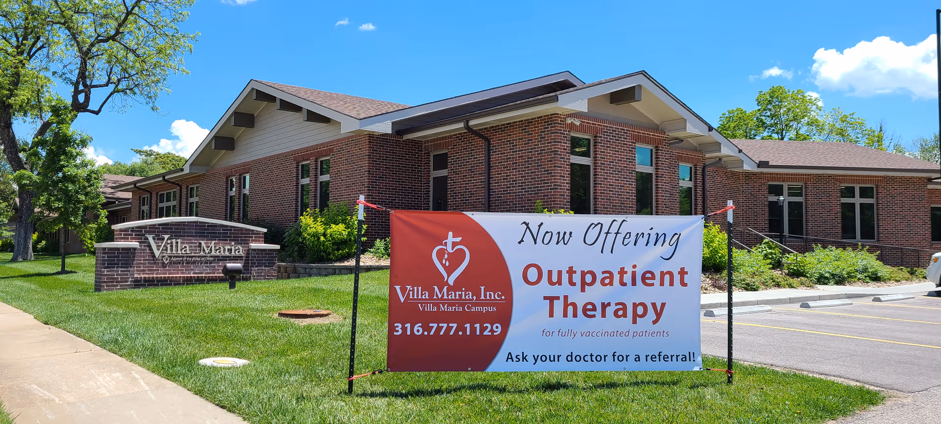 Exterior view of Villa Maria building with a brick facade under a clear blue sky. A large sign in front of the building advertises outpatient therapy for fully vaccinated patients, including contact information. The surrounding area has green grass, trees, and a parking lot.