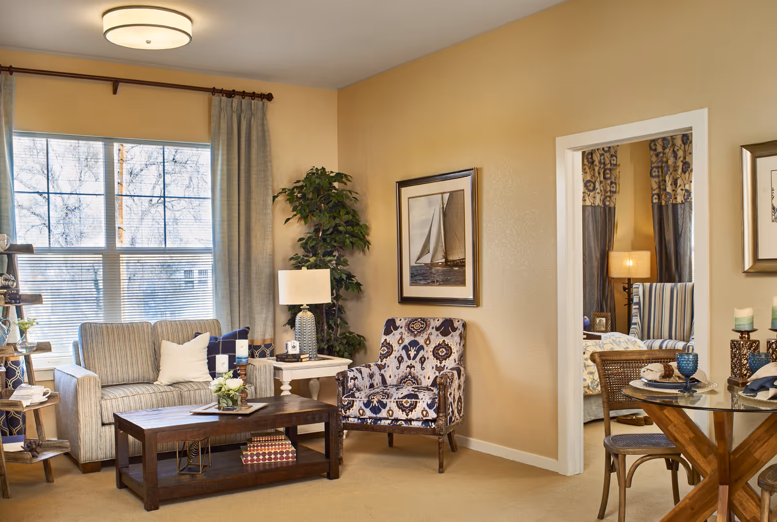 A cozy living room area with a striped sofa, a patterned armchair, a wooden coffee table with decorative items, a side table with a lamp, and a large window with curtains. Adjacent to the living room is a dining area with a glass-top table and wooden chairs, and a glimpse into another room with additional seating and a lamp.