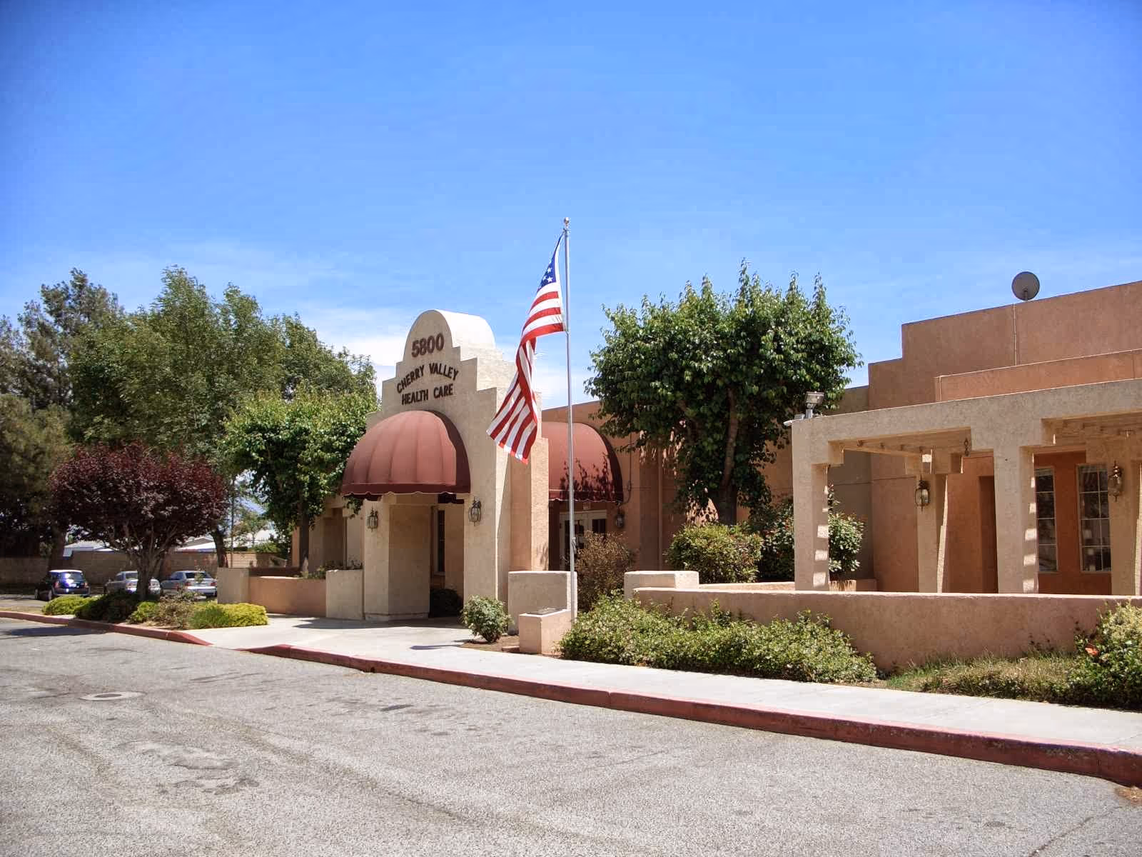 Exterior view of Cherry Valley Health Care building with beige walls, a maroon awning over the entrance, an American flag on a flagpole, and surrounding trees and shrubs under a clear blue sky.